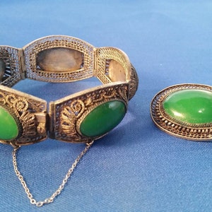 May include: A silver-toned filigree bracelet with oval green jade stones and a matching oval brooch with a green jade stone, set against a blue background. The bracelet has a safety chain.