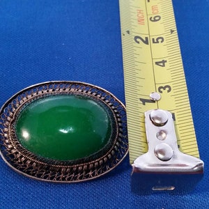 May include: Vintage jewelry set featuring a green oval stone set in a filigree metal frame. The brooch measures approximately 1.5 inches in length. The set includes a matching bracelet. The background is a solid blue.