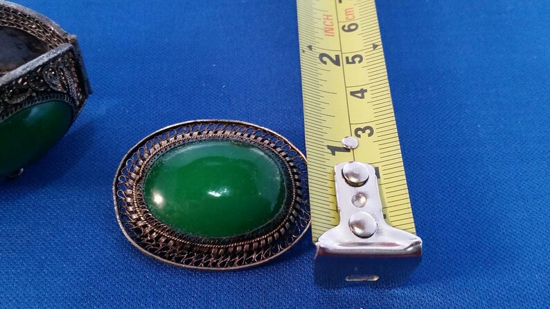 May include: Vintage jewelry set featuring a green oval stone set in a filigree metal frame. The brooch measures approximately 1.5 inches in length. The set includes a matching bracelet. The background is a solid blue.