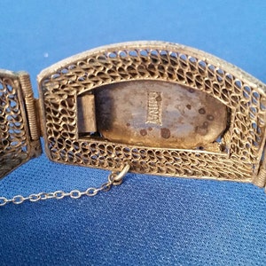 May include: Close-up of a vintage silver filigree bracelet with a rectangular clasp marked "CHINA". The bracelet features intricate openwork designs and a small safety chain. A green stone is visible on the right.