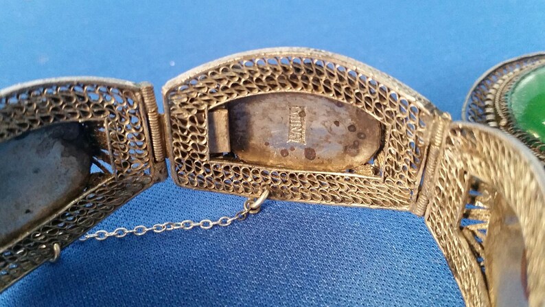 May include: Close-up of a vintage silver filigree bracelet with a rectangular clasp marked "CHINA". The bracelet features intricate openwork designs and a small safety chain. A green stone is visible on the right.