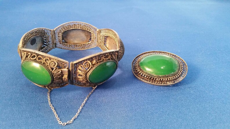 May include: A silver-toned filigree bracelet with oval green jade stones and a matching oval brooch with a green jade stone, set against a blue background. The bracelet has a safety chain.