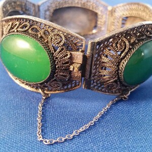 May include: An antique silver-toned filigree bracelet with two large oval green stones. The bracelet is open, revealing the clasp and a small chain. A matching oval green stone earring is also visible.