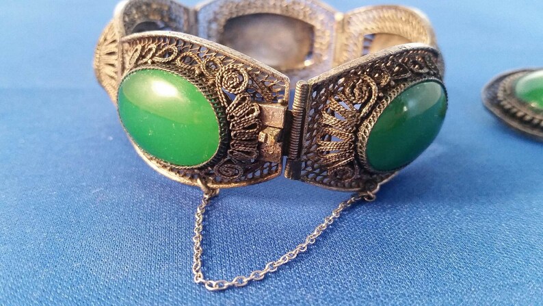 May include: An antique silver-toned filigree bracelet with two large oval green stones. The bracelet is open, revealing the clasp and a small chain. A matching oval green stone earring is also visible.