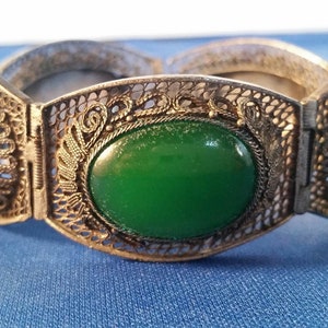 May include: An antique silver-toned bracelet with filigree detailing and large oval green stones. The bracelet is hinged and has a matching green stone accent piece. The jewelry is displayed on a blue surface.