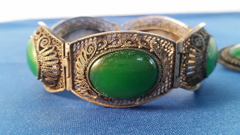 May include: An antique silver-toned bracelet with filigree detailing and large oval green stones. The bracelet is hinged and has a matching green stone accent piece. The jewelry is displayed on a blue surface.