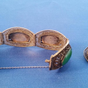 May include: A silver filigree bracelet with oval green jade stones and a matching oval jade brooch. The bracelet is partially open, revealing the intricate metalwork and the clasp. The jewelry is displayed on a blue surface.