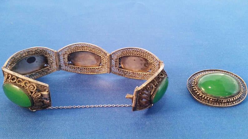 May include: A silver filigree bracelet with oval green jade stones and a matching oval jade brooch. The bracelet is partially open, revealing the intricate metalwork and the clasp. The jewelry is displayed on a blue surface.
