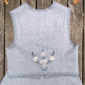 K&ouml;nnte beinhalten: Eine graue Wollweste mit einem floralen Stickmuster auf der R&uuml;ckseite. Die Weste hat einen geh&auml;kelten Saum an der Unterseite und den Arml&ouml;chern.