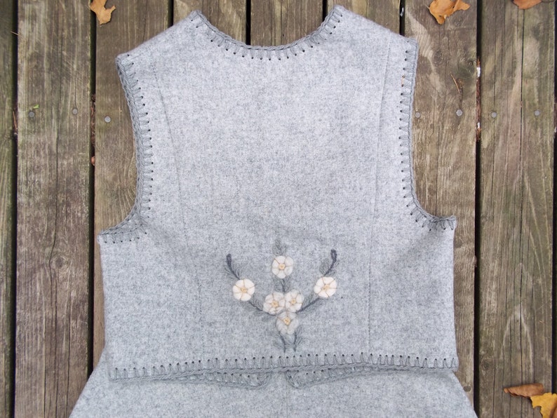 K&ouml;nnte beinhalten: Eine graue Wollweste mit einem floralen Stickmuster auf der R&uuml;ckseite. Die Weste hat einen geh&auml;kelten Saum an der Unterseite und den Arml&ouml;chern.