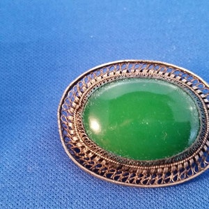 May include: An oval-shaped green stone brooch with a filigree metal frame. The brooch has a bright green center and a detailed, openwork border. The metal has a silver or antique finish. The background is a solid blue.