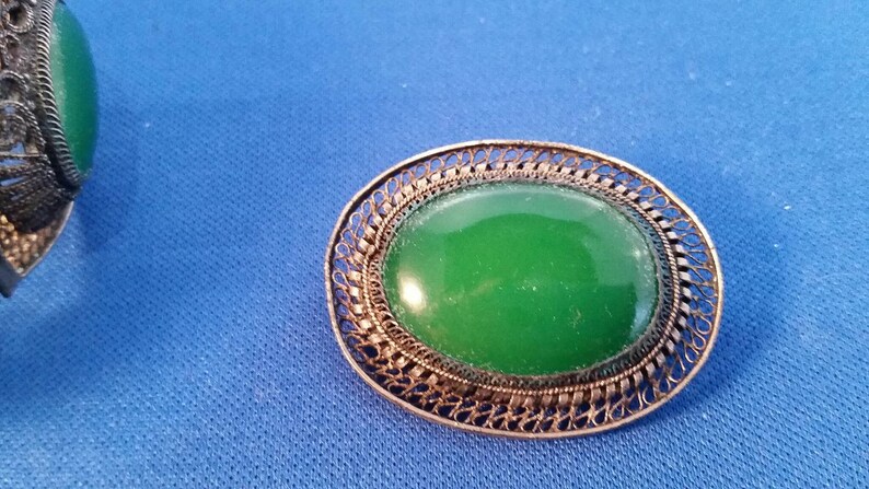 May include: An oval-shaped green stone brooch with a filigree metal frame. The brooch has a bright green center and a detailed, openwork border. The metal has a silver or antique finish. The background is a solid blue.