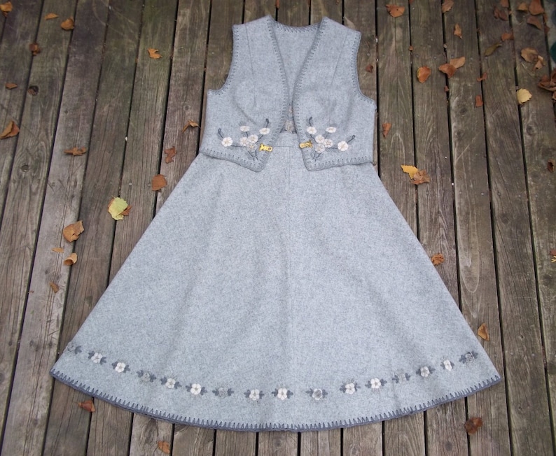 K&ouml;nnte beinhalten: Ein graues Wollkleid mit einer passenden Weste. Beide Teile sind mit gestickten Blumenmotiven in Wei&szlig; verziert. Das Kleid hat einen vollen Rock und ein tailliertes Oberteil.
