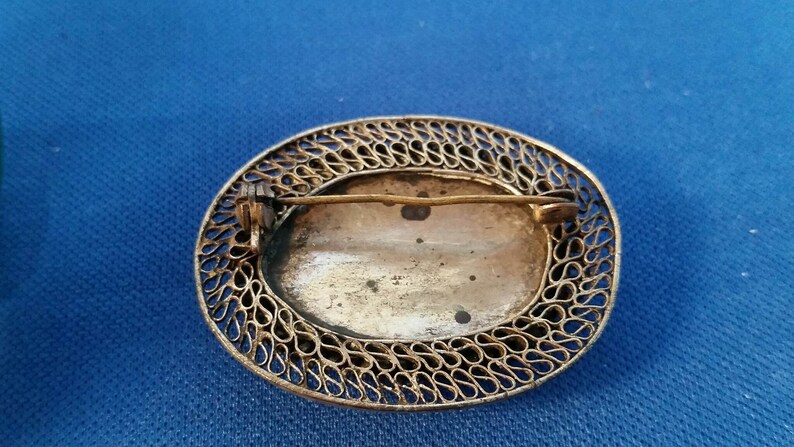 May include: An antique oval brooch with a filigree border. The brooch has a pin on the back for attachment. The center of the brooch has a faded image. The metal has a tarnished appearance.
