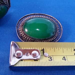 May include: An oval green gemstone brooch with an ornate filigree metal frame. The brooch is approximately 2 inches long. A matching bracelet with a similar green stone is visible. The items are on a blue surface.