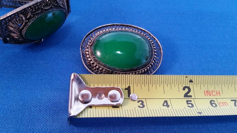 May include: An oval green gemstone brooch with an ornate filigree metal frame. The brooch is approximately 2 inches long. A matching bracelet with a similar green stone is visible. The items are on a blue surface.