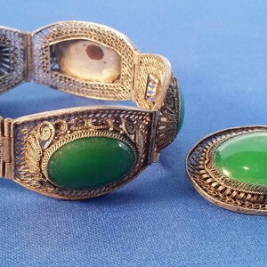May include: A vintage jewelry set featuring a filigree bracelet and a matching brooch. The bracelet has six oval green stones and intricate metalwork. The brooch has a single oval green stone. Both pieces are set against a blue background.