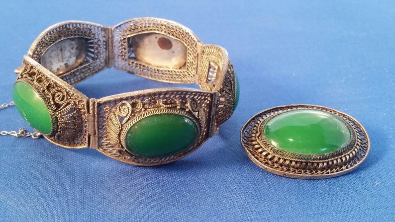 May include: A vintage jewelry set featuring a filigree bracelet and a matching brooch. The bracelet has six oval green stones and intricate metalwork. The brooch has a single oval green stone. Both pieces are set against a blue background.