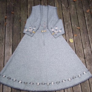 K&ouml;nnte beinhalten: Ein graues Wollkleid mit einer passenden Weste. Das Kleid und die Weste sind mit wei&szlig;en Blumen bestickt und haben einen grauen Saum.
