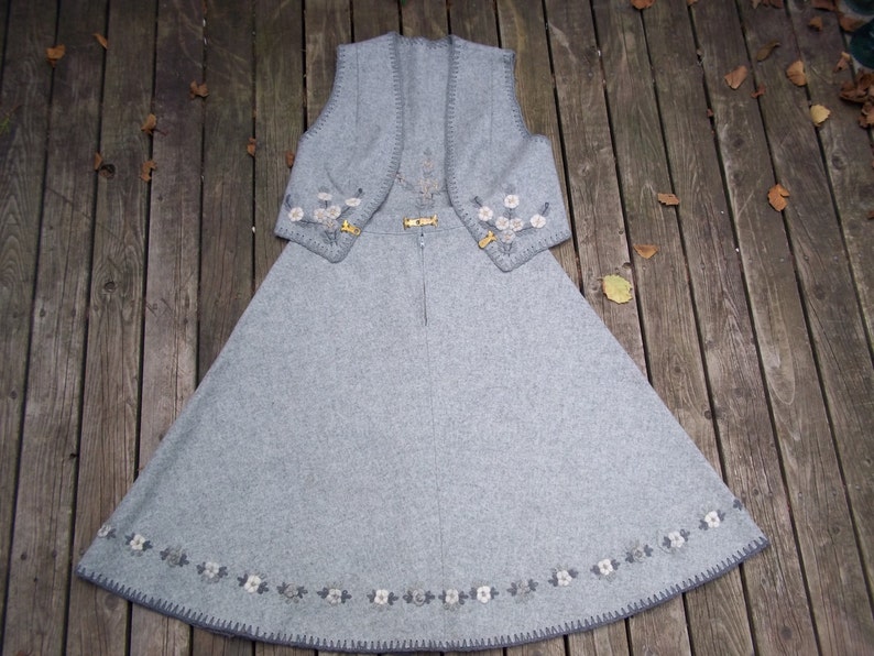 K&ouml;nnte beinhalten: Ein graues Wollkleid mit einer passenden Weste. Das Kleid und die Weste sind mit wei&szlig;en Blumen bestickt und haben einen grauen Saum.