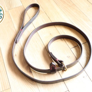 May include: A brown leather dog leash with a brass clasp and red stitching. The leash is coiled on a wooden surface.