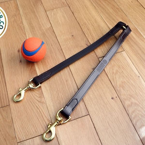 May include: A brown leather dog leash with two gold metal clips. The leash is lying on a wooden floor with a blue and orange ball nearby.