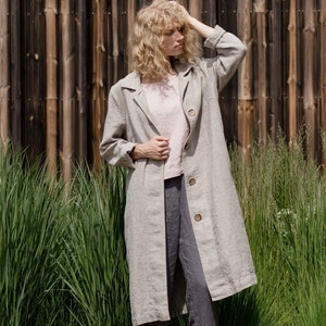 Può includere: Una donna indossa un lungo cappotto di lino grigio chiaro con chiusura a bottoni. È in piedi davanti a una recinzione di legno e erba verde.