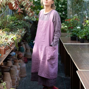 Peut inclure: Une femme portant un long tablier en lin rose se tient dans une serre. Elle est entourée de plantes en pot et d'étagères de fournitures de jardinage.