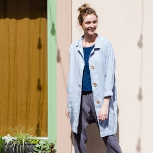 Peut inclure: Une femme portant une veste en lin bleu clair sur une chemise bleu foncé et un pantalon gris. Elle se tient devant un bâtiment avec une fenêtre et un mur brun clair.