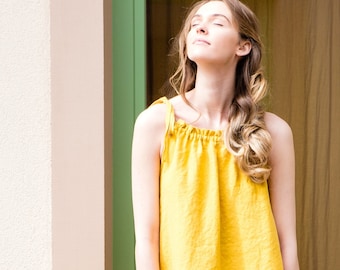 May include: A woman wearing a bright yellow sleeveless linen dress with a gathered neckline. She is standing in front of a doorway with her eyes closed and head tilted back.