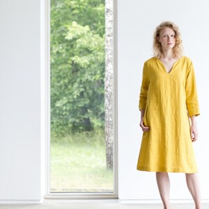 May include: A woman wearing a yellow linen dress with a V-neck and 3/4 sleeves. She is standing in front of a window with a view of trees.