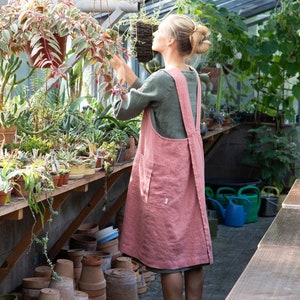 以下が含まれることがあります： 温室の中で植物を見ている、大きなポケットが付いたピンクのリネンエプロンを着た女性