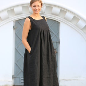 May include: A woman wearing a black sleeveless linen dress with pockets. She is standing on a stone surface in front of a white building with a gray door.