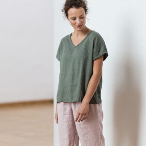 May include: A woman is wearing a green linen top with a v-neck and short sleeves. She is also wearing pink linen shorts.