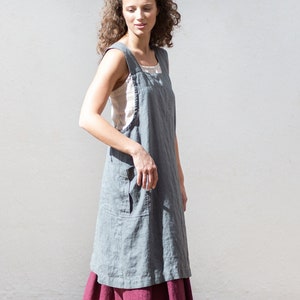 Könnte beinhalten: Ein graues Leinen-Schürzenkleid mit quadratischem Ausschnitt und großer Fronttasche. Das Kleid ist über einem bordeauxfarbenen Rock geschichtet. Das Model trägt olivgrüne Wildlederschuhe.
