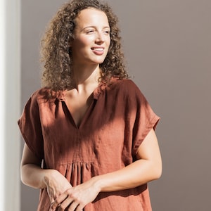 May include: A woman with curly brown hair is wearing a rust-colored linen dress with a v-neck and short sleeves. She is standing in front of a white wall and looking to the right.