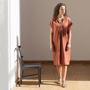 May include: A woman wearing a terracotta linen dress with a v-neck and short sleeves stands in front of a white wall. She is wearing sandals and looking to the side.