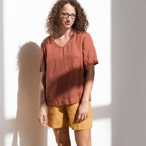 May include: A woman wearing a rust-colored linen top with a v-neck and a pair of mustard yellow linen shorts.
