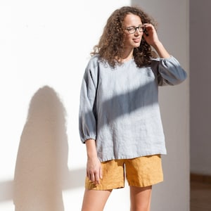May include: A woman wearing a light blue linen top with a relaxed fit and a pair of yellow linen shorts. She is wearing glasses and has long brown hair.