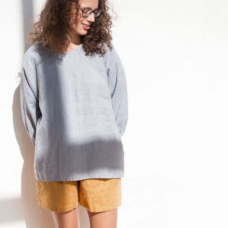 May include: A woman wearing a light blue linen shirt and mustard yellow linen shorts. She is wearing glasses and has curly brown hair.