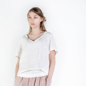 May include: A white linen top with a v-neck and short sleeves. The model is wearing a light pink linen skirt.