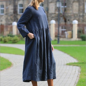 May include: A woman wearing a long, dark blue linen dress with a gathered waist and long sleeves. She is wearing brown lace-up boots.