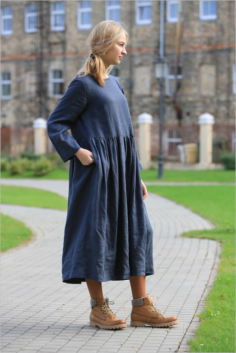 May include: A woman wearing a long, dark blue linen dress with a gathered waist and long sleeves. She is wearing brown lace-up boots.