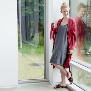 May include: A woman wearing a red linen robe over a gray linen dress. She is standing near a window and looking to the side.