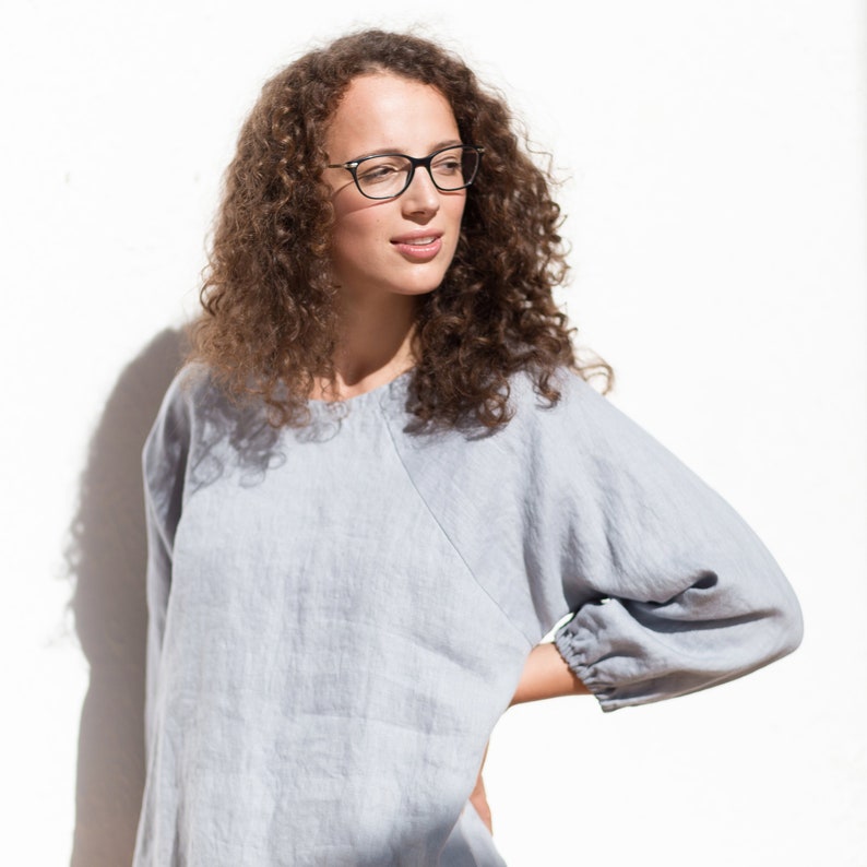 May include: A woman with curly brown hair wearing black framed glasses and a light blue linen top.