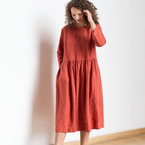 May include: A woman wearing a rust-colored linen dress with long sleeves and a gathered skirt. She is standing on a wooden floor with a white wall behind her.