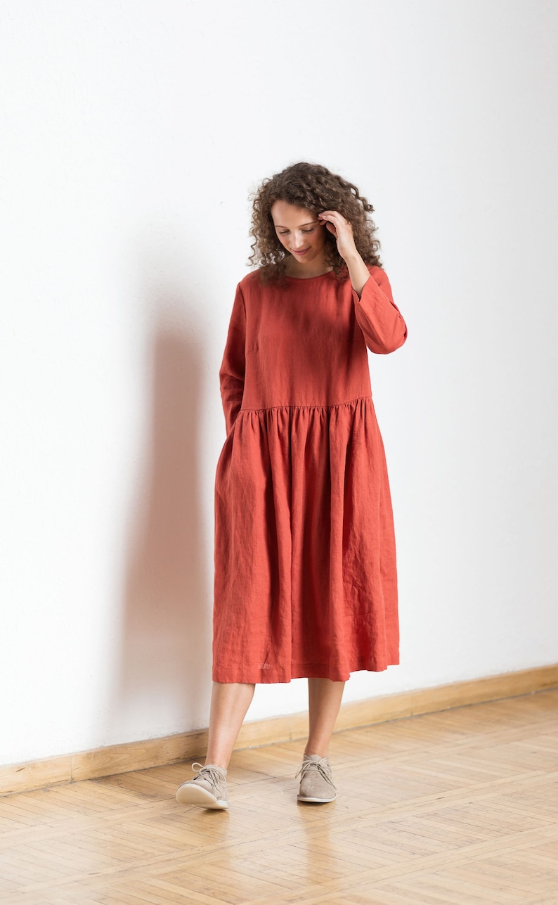 May include: A woman wearing a rust-colored linen dress with long sleeves and a gathered skirt. She is standing on a wooden floor with a white wall behind her.
