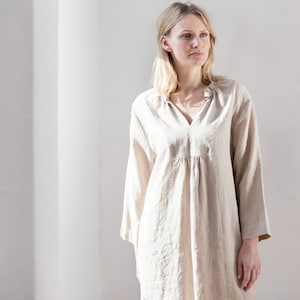 May include: A woman wearing a light beige linen dress with a V-neck and long sleeves. The dress has a gathered waist and a relaxed fit.