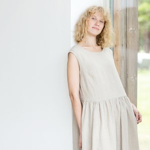 May include: A woman wearing a light beige linen dress with a gathered skirt. She is standing next to a window.