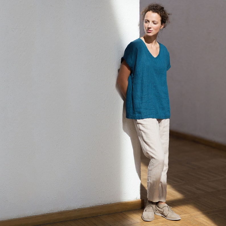 May include: A woman wearing a teal blue linen top with a v-neck and a pair of beige linen pants. She is standing next to a white wall.